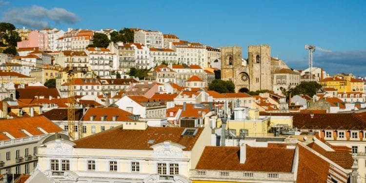 Una vista panoramica di una città europea con un mix di edifici colorati con tetti di tegole rosse. Sullo sfondo è visibile un'importante struttura storica in pietra con torri gemelle, che evoca pensieri di Investimenti stranieri immobili Lisbona. Il cielo è limpido e azzurro. I paesaggi urbani e collinari si fondono perfettamente.