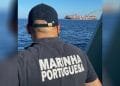 Una persona in uniforme della marina con la scritta "MARINHA PORTUGUESA" sulla schiena guarda il mare da una barca. La scena è cupa, forse legata ad una ragazza scomparsa in mare. In lontananza, sull'acqua è visibile una grande nave mercantile con la scritta "MSC" sulla fiancata, che emette una scia di fumo.