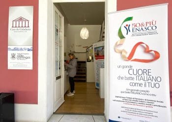 Una persona si trova all'interno di un edificio in prossimità di un ingresso, interagendo con una bacheca. A destra, un grande striscione riporta informazioni su 50&PIÙ ENASCO, istituto italiano di assistenza sociale. Sulla parete laterale sinistra sono presenti le indicazioni per Casa da Cidadania e Junta de Freguesia Sao Domingos, parte della sede patronato di Lisbona.