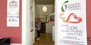 Una persona si trova all'interno di un edificio in prossimità di un ingresso, interagendo con una bacheca. A destra, un grande striscione riporta informazioni su 50&PIÙ ENASCO, istituto italiano di assistenza sociale. Sulla parete laterale sinistra sono presenti le indicazioni per Casa da Cidadania e Junta de Freguesia Sao Domingos, parte della sede patronato di Lisbona.