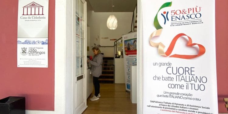 Una persona si trova all'interno di un edificio in prossimità di un ingresso, interagendo con una bacheca. A destra, un grande striscione riporta informazioni su 50&PIÙ ENASCO, istituto italiano di assistenza sociale. Sulla parete laterale sinistra sono presenti le indicazioni per Casa da Cidadania e Junta de Freguesia Sao Domingos, parte della sede patronato di Lisbona.