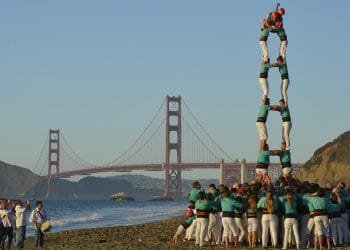 Un folto gruppo di persone forma una torre umana, che ricorda le tradizionali torri umane viste in Catalogna, su una spiaggia con l'iconico Golden Gate Bridge sullo sfondo. Diversi individui sostengono la base mentre altri si arrampicano verso l'alto per creare più livelli, raggiungendo altezze impressionanti sotto un cielo limpido.