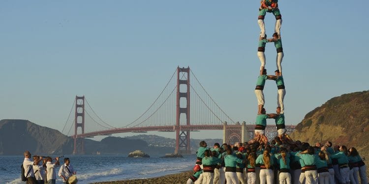 Un folto gruppo di persone forma una torre umana, che ricorda le tradizionali torri umane viste in Catalogna, su una spiaggia con l'iconico Golden Gate Bridge sullo sfondo. Diversi individui sostengono la base mentre altri si arrampicano verso l'alto per creare più livelli, raggiungendo altezze impressionanti sotto un cielo limpido.