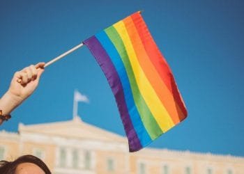 Una mano che tiene una bandiera arcobaleno contro un cielo azzurro chiaro alla Pride Parade LGBTQI+ dell'Algarve. Sullo sfondo è visibile un grande edificio con una bandiera che sventola in alto. La bandiera arcobaleno simboleggia l’orgoglio LGBTQ+.