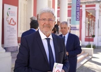 Un uomo con i capelli grigi e gli occhiali, Antonio Paoletti Enasco, vestito con abito scuro e cravatta, sta all'aperto con in mano un libretto. Altre persone in giacca e cravatta sono sullo sfondo vicino ad un edificio con colonne bianche e uno striscione con la scritta "CASA DA CIDADANIA.
