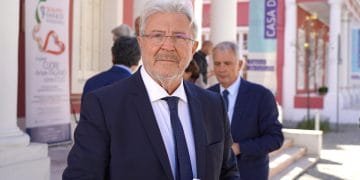 Un uomo con i capelli grigi e gli occhiali, Antonio Paoletti Enasco, vestito con abito scuro e cravatta, sta all'aperto con in mano un libretto. Altre persone in giacca e cravatta sono sullo sfondo vicino ad un edificio con colonne bianche e uno striscione con la scritta "CASA DA CIDADANIA.