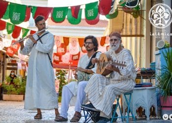 Tre musicisti vestiti con abiti tradizionali si esibiscono in una strada acciottolata, con stendardi festivi appesi sopra. Uno suona il flauto, l'altro tiene in mano un tamburo e il terzo strimpella uno strumento a corde. L'ambientazione sembra essere il Festival Islamico Mértola, che mette in mostra vivaci celebrazioni culturali.