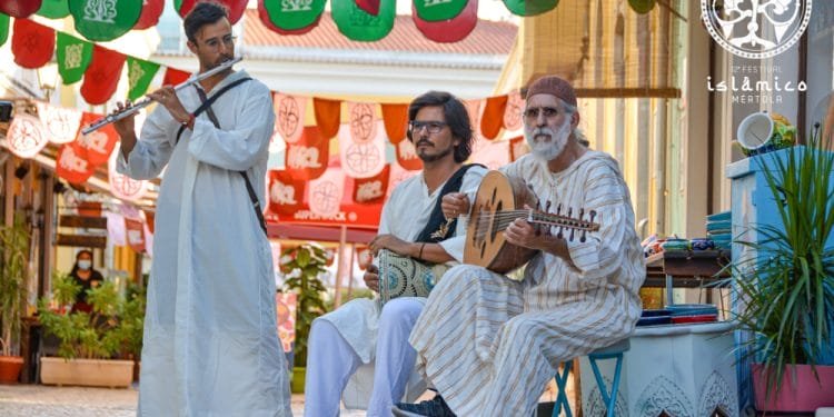 Tre musicisti vestiti con abiti tradizionali si esibiscono in una strada acciottolata, con stendardi festivi appesi sopra. Uno suona il flauto, l'altro tiene in mano un tamburo e il terzo strimpella uno strumento a corde. L'ambientazione sembra essere il Festival Islamico Mértola, che mette in mostra vivaci celebrazioni culturali.