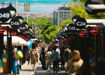 Un affollato mercato all'aperto, parte della Fiera Libro Lisbona, è caratterizzato da bancarelle nere contrassegnate da numeri bianchi; la gente passeggia tra le bancarelle nelle giornate di sole, con alberi e vista sul mare e sugli edifici della città sullo sfondo.