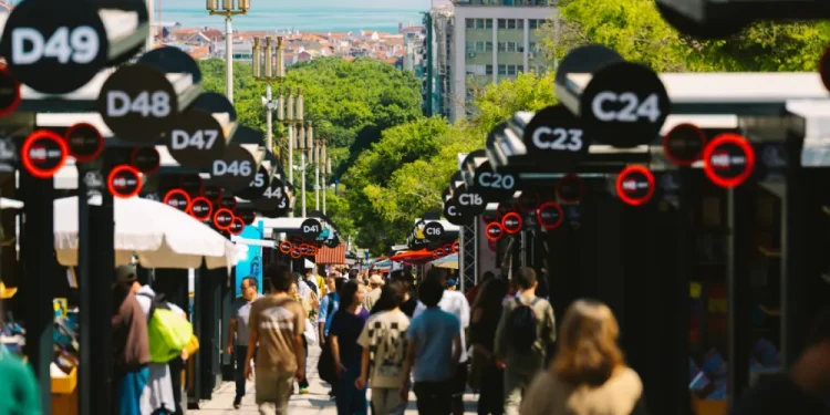 Un affollato mercato all'aperto, parte della Fiera Libro Lisbona, è caratterizzato da bancarelle nere contrassegnate da numeri bianchi; la gente passeggia tra le bancarelle nelle giornate di sole, con alberi e vista sul mare e sugli edifici della città sullo sfondo.
