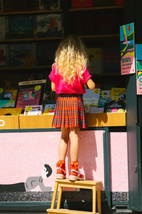 Una bambina di spalle su uno sgabello cerca alla fiera di Lisbona un libro da alcuni contenitori.