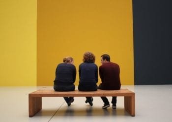 Tre persone si siedono e conversano su una panca di legno in una stanza spaziosa e minimalista con pareti bicolore giallo e grigio scuro. Visti di spalle creano una scena contemplativa, che ricorda la tranquillità celebrata nella Giornata Internazionale dei Musei.