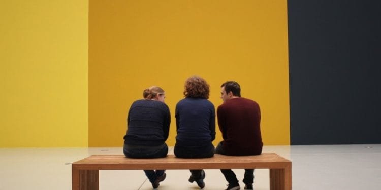 Tre persone si siedono e conversano su una panca di legno in una stanza spaziosa e minimalista con pareti bicolore giallo e grigio scuro. Visti di spalle creano una scena contemplativa, che ricorda la tranquillità celebrata nella Giornata Internazionale dei Musei.