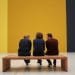 Tre persone si siedono e conversano su una panca di legno in una stanza spaziosa e minimalista con pareti bicolore giallo e grigio scuro. Visti di spalle creano una scena contemplativa, che ricorda la tranquillità celebrata nella Giornata Internazionale dei Musei.