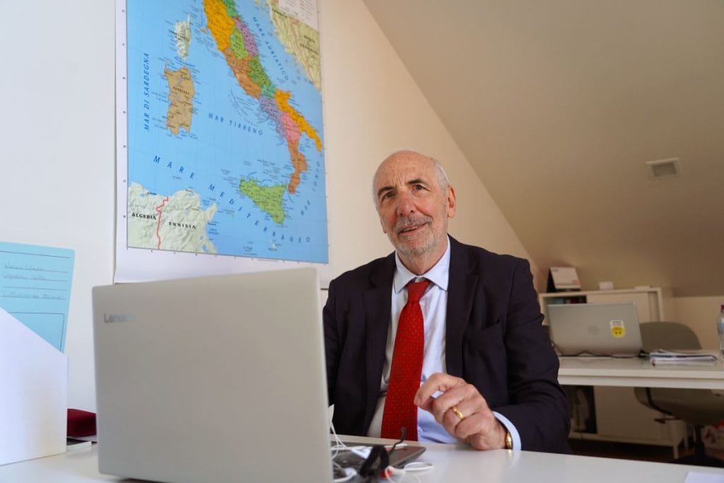 Un uomo anziano con la barba, che indossa un abito e una cravatta rossa, siede alla scrivania e lavora su un laptop Lenovo. Dietro di lui c'è una mappa dell'Italia appesa al muro. La stanza sembra essere un ufficio con il soffitto inclinato. Questa potrebbe facilmente essere la routine quotidiana di Guido Moretti, profondamente assorbito dai suoi compiti.