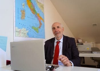 Un uomo anziano con la barba, che indossa un abito e una cravatta rossa, siede alla scrivania e lavora su un laptop Lenovo. Dietro di lui c'è una mappa dell'Italia appesa al muro. La stanza sembra essere un ufficio con il soffitto inclinato. Questa potrebbe facilmente essere la routine quotidiana di Guido Moretti, profondamente assorbito dai suoi compiti.