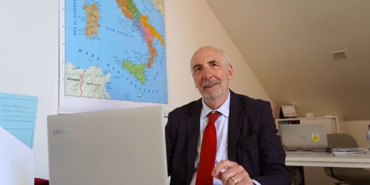 Un uomo anziano con la barba, che indossa un abito e una cravatta rossa, siede alla scrivania e lavora su un laptop Lenovo. Dietro di lui c'è una mappa dell'Italia appesa al muro. La stanza sembra essere un ufficio con il soffitto inclinato. Questa potrebbe facilmente essere la routine quotidiana di Guido Moretti, profondamente assorbito dai suoi compiti.