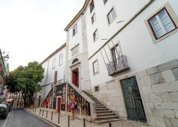 Una scena di strada a Lisbona, in Portogallo, presenta un classico edificio bianco con gradini in pietra decorati e decorati da vivaci murales. Alcune persone camminano o stanno in piedi vicino alla struttura aperta. La strada acciottolata, fiancheggiata da edifici e alberi, si estende in lontananza, evocando immagini di antichi monasteri.