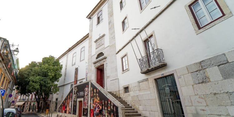 Una scena di strada a Lisbona, in Portogallo, presenta un classico edificio bianco con gradini in pietra decorati e decorati da vivaci murales. Alcune persone camminano o stanno in piedi vicino alla struttura aperta. La strada acciottolata, fiancheggiata da edifici e alberi, si estende in lontananza, evocando immagini di antichi monasteri.