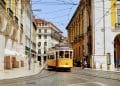 Un tram giallo percorre una strada acciottolata fiancheggiata da edifici storici e archi in Portogallo. Si vedono pedoni camminare e stare sui marciapiedi in una giornata di sole. La scena cattura una miscela di architettura del vecchio mondo e transito urbano, rendendolo uno dei migliori paesi eventi.