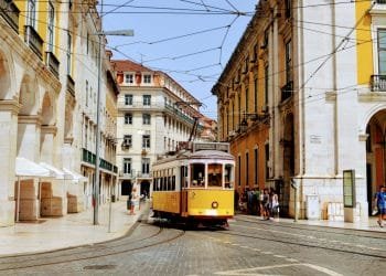 Un tram giallo percorre una strada acciottolata fiancheggiata da edifici storici e archi in Portogallo. Si vedono pedoni camminare e stare sui marciapiedi in una giornata di sole. La scena cattura una miscela di architettura del vecchio mondo e transito urbano, rendendolo uno dei migliori paesi eventi.