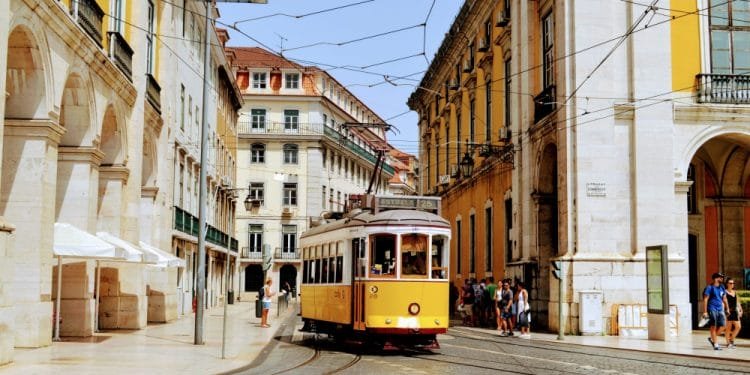 Un tram giallo percorre una strada acciottolata fiancheggiata da edifici storici e archi in Portogallo. Si vedono pedoni camminare e stare sui marciapiedi in una giornata di sole. La scena cattura una miscela di architettura del vecchio mondo e transito urbano, rendendolo uno dei migliori paesi eventi.