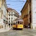 Un tram giallo percorre una strada acciottolata fiancheggiata da edifici storici e archi in Portogallo. Si vedono pedoni camminare e stare sui marciapiedi in una giornata di sole. La scena cattura una miscela di architettura del vecchio mondo e transito urbano, rendendolo uno dei migliori paesi eventi.