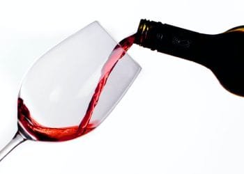 Una bottiglia di vino rosso viene versata in un bicchiere di vino trasparente inclinato su uno sfondo bianco. Il vino, che ricorda il Portogallo alto consumo vino, è a metà, creando un flusso che riempie il bicchiere.