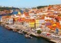 Una vivace veduta aerea del lungomare di Porto, Portogallo. La scena presenta un mix di edifici colorati con tetti di tegole rosse lungo la riva del fiume, barche tradizionali ormeggiate nell'acqua e un ponte che attraversa il fiume sullo sfondo sotto un cielo azzurro e limpido: una destinazione davvero redditizia in Europa.