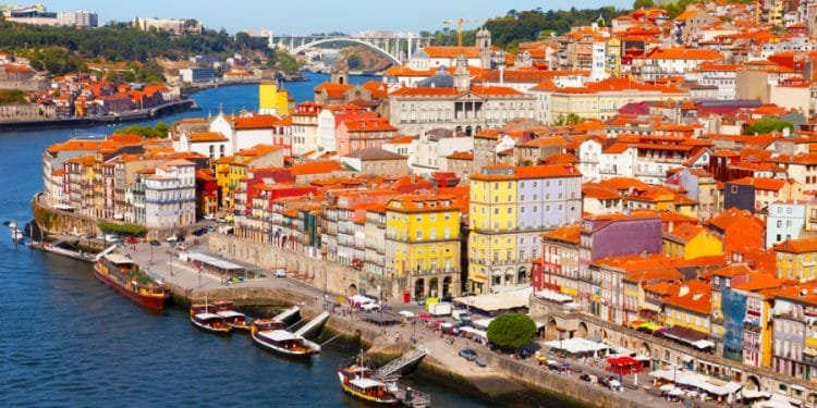 Una vivace veduta aerea del lungomare di Porto, Portogallo. La scena presenta un mix di edifici colorati con tetti di tegole rosse lungo la riva del fiume, barche tradizionali ormeggiate nell'acqua e un ponte che attraversa il fiume sullo sfondo sotto un cielo azzurro e limpido: una destinazione davvero redditizia in Europa.