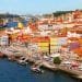 Una vivace veduta aerea del lungomare di Porto, Portogallo. La scena presenta un mix di edifici colorati con tetti di tegole rosse lungo la riva del fiume, barche tradizionali ormeggiate nell'acqua e un ponte che attraversa il fiume sullo sfondo sotto un cielo azzurro e limpido: una destinazione davvero redditizia in Europa.
