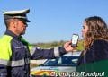 Un agente di polizia che indossa una giacca riflettente e un berretto con visiera tiene in mano un dispositivo per l'etilometro davanti a una donna. Sullo sfondo è visibile un'auto della polizia. Il testo sull'immagine recita "Operação Roadpol Álcool e Drogas", evidenziando la campagna polizia alcol guida, e include i loghi di GNR e Roadpol.