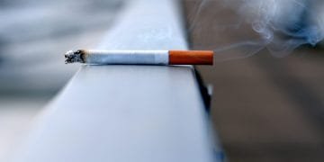 Una sigaretta accesa con il fumo che sale dalla punta poggia su una superficie metallica. L'immagine è focalizzata sulla sigaretta, mentre lo sfondo è sfocato, enfatizzando la sigaretta e il fumo, evidenziando per tutti gli occhi un chiaro segno di *divieto fumare*.