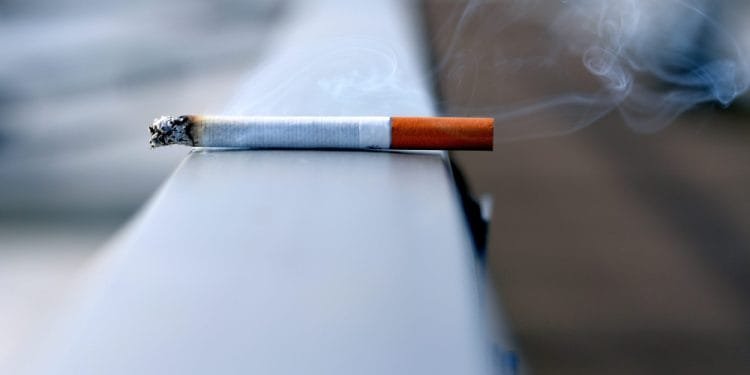 Una sigaretta accesa con il fumo che sale dalla punta poggia su una superficie metallica. L'immagine è focalizzata sulla sigaretta, mentre lo sfondo è sfocato, enfatizzando la sigaretta e il fumo, evidenziando per tutti gli occhi un chiaro segno di *divieto fumare*.