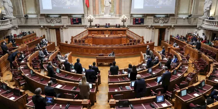 Camera legislativa con numerosi posti disposti a semicerchio. Diverse persone stanno in piedi e conversano nella stanza, probabilmente discutendo di recenti decisioni come quando la legge eutanasia è stata approvata in Portogallo. Sullo sfondo sono visibili grandi schermi e statue, insieme a un prominente podio per oratori nella parte anteriore.