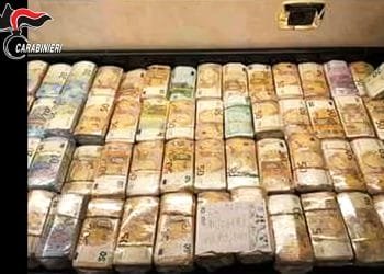 I pacchi di banconote in euro sono impilati ordinatamente in uno spazio di archiviazione. Le banconote sono prevalentemente banconote da 50€. Inoltre, nell'angolo in alto a sinistra è presente l'emblema dei "Carabinieri", che suggerisce un collegamento con le forze dell'ordine e potenzialmente allude a un'operazione legata alla Ndrangheta in Portogallo.