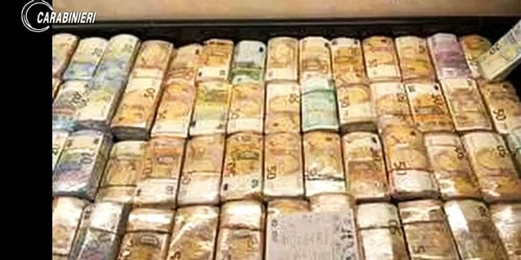 I pacchi di banconote in euro sono impilati ordinatamente in uno spazio di archiviazione. Le banconote sono prevalentemente banconote da 50€. Inoltre, nell'angolo in alto a sinistra è presente l'emblema dei "Carabinieri", che suggerisce un collegamento con le forze dell'ordine e potenzialmente allude a un'operazione legata alla Ndrangheta in Portogallo.