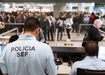 Due agenti in uniforme, con la scritta "Polícia SEF" sulle magliette, sono seduti alle scrivanie e gestiscono una coda di passeggeri in un aeroporto affollato. Nonostante i reclami contro SEF Portogallo, la scena rimane affollata di persone in fila, sfocate sullo sfondo.