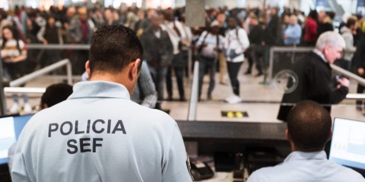 Due agenti in uniforme, con la scritta "Polícia SEF" sulle magliette, sono seduti alle scrivanie e gestiscono una coda di passeggeri in un aeroporto affollato. Nonostante i reclami contro SEF Portogallo, la scena rimane affollata di persone in fila, sfocate sullo sfondo.