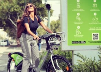 Una donna con gli occhiali da sole e una camicia nera sorride mentre guida una bicicletta verde a noleggio in una giornata di sole, godendosi l'ampia rete GIRA. Porta uno zaino marrone. Dietro di lei c'è un pannello informativo verde con testo e icone, e sullo sfondo sono visibili gli alberi, tipico del fascino di Lisbona per i residenti.