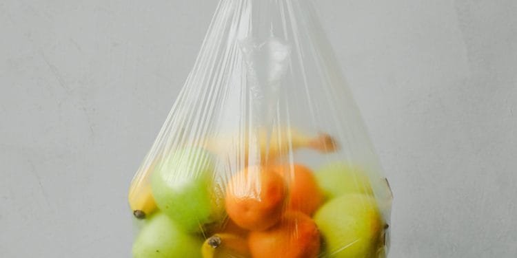 Un sacchetto di plastica trasparente pieno di frutta assortita, tra cui mele verdi e gialle, arance e una banana, è sospeso su un semplice sfondo grigio chiaro. I colori vivaci dei frutti sono visibili attraverso la plastica, evidenziando la necessità di prendere in considerazione iniziative di divieto sacchetti plastica.