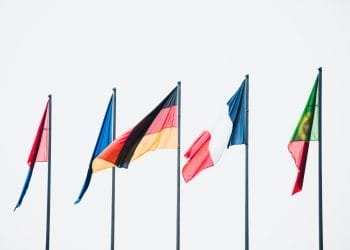 Cinque bandiere nazionali sventolano sui pennoni contro un cielo bianco e semplice. Da sinistra a destra, le bandiere sono rossa, blu e gialla, nera-rossa-oro (Germania), blu-bianca-rossa (Francia) e verde-rossa con stemma giallo (Portogallo), a simboleggiare la sua ricca storia e la sistema fiscale portoghese complesso.