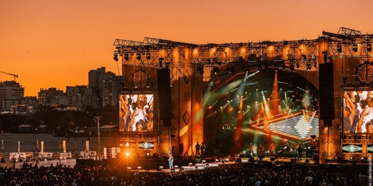 Una grande folla si riunisce per l'Afro Nation festival su una spiaggia al tramonto. Il palco è ben illuminato con luci colorate e due grandi schermi su entrambi i lati mostrano gli artisti che si divertono a ritmo di afrobeat. Il cielo è di una tonalità arancione e gli edifici sono visibili sullo sfondo.