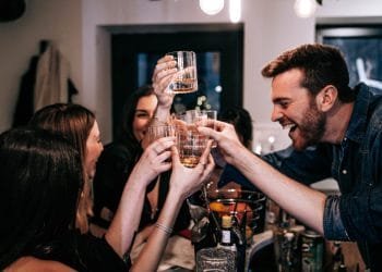 Un gruppo di persone tiene in mano i bicchieri in un brindisi mentre sorride e ride. Si riuniscono attorno a un tavolo con bevande e snack, creando un'atmosfera vivace e festosa. L'ambientazione sembra essere al chiuso, forse in un Airbnb in Portogallo, nonostante il divieto di feste.