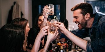 Un gruppo di persone tiene in mano i bicchieri in un brindisi mentre sorride e ride. Si riuniscono attorno a un tavolo con bevande e snack, creando un'atmosfera vivace e festosa. L'ambientazione sembra essere al chiuso, forse in un Airbnb in Portogallo, nonostante il divieto di feste.