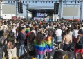 Una grande folla si raduna all'aperto davanti al palco etichettato "Liberty" all'Arraial Lisboa Pride. Molte persone sono avvolte in bandiere arcobaleno, che celebrano l’orgoglio LGBTQ+. La scena presenta individui diversi e un'atmosfera festosa con cieli sereni sopra l'Arraial Lisboa Pride.