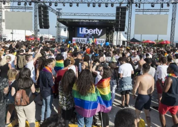 Una grande folla si raduna all'aperto davanti al palco etichettato "Liberty" all'Arraial Lisboa Pride. Molte persone sono avvolte in bandiere arcobaleno, che celebrano l’orgoglio LGBTQ+. La scena presenta individui diversi e un'atmosfera festosa con cieli sereni sopra l'Arraial Lisboa Pride.