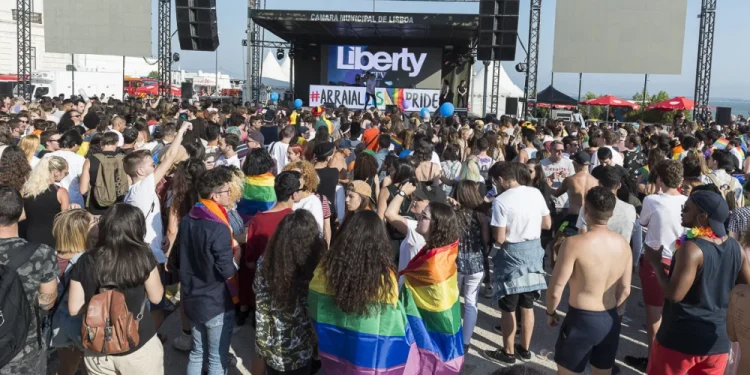 Una grande folla si raduna all'aperto davanti al palco etichettato "Liberty" all'Arraial Lisboa Pride. Molte persone sono avvolte in bandiere arcobaleno, che celebrano l’orgoglio LGBTQ+. La scena presenta individui diversi e un'atmosfera festosa con cieli sereni sopra l'Arraial Lisboa Pride.
