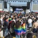 Una grande folla si raduna all'aperto davanti al palco etichettato "Liberty" all'Arraial Lisboa Pride. Molte persone sono avvolte in bandiere arcobaleno, che celebrano l’orgoglio LGBTQ+. La scena presenta individui diversi e un'atmosfera festosa con cieli sereni sopra l'Arraial Lisboa Pride.