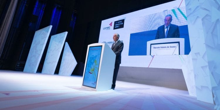 Un uomo sta dietro un podio su un grande palco ben illuminato. Un grande schermo accanto a lui mostra la sua immagine e il testo "Marcelo Rebelo de Sousa". Lo sfondo presenta pannelli astratti bianchi e loghi per "COTEC Portogallo" e "Portugal Covid-19 Response", evidenziando le collaborazioni con Italia e Spagna.